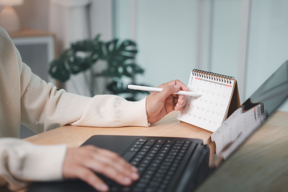 Worker planning to write paper work report presentation meeting dates on calendar, using laptop computer at wooden desk in office, organizing timeline for corporate business project Salaries eloignes